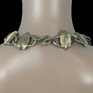May include: A necklace featuring a series of textured, gold-toned leaf-shaped metal links connected by smaller silver-toned rings. The piece includes a central hook-and-eye clasp mechanism that secures the chain around the neck area.