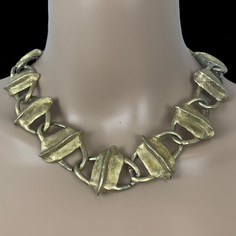 May include: A gold-toned statement necklace featuring a series of interconnected, textured, leaf-shaped metal links. Each link has a raised central ridge and a brushed, matte finish, creating a continuous chain design that rests around the neck.