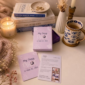 Puede incluir: Una baraja de My Cozy Tarot en una caja morada clara con cartas a juego, una taza blanca con patrones de ojos azules, una pila de libros incluyendo The Little Book of Hygge, una vela en un frasco de vidrio, un cuenco decorativo y flores secas.