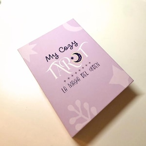 Op de afbeelding: Een lila tarotkaartdoos met de tekst "My Cozy Tarot" in een speels lettertype. Het woord "Tarot" heeft een maan- en sterontwerp. De tekst "La Magia Del Orden" staat eronder, met ster- en bloemaccenten.