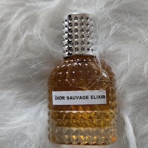 Może przedstawiać: Mała butelka perfum Dior Sauvage Elixir. Butelka ma bursztynowy kolor ze srebrną, teksturowaną nakrętką. Etykieta zawiera napis "DIOR SAUVAGE ELIXIR" w białym tekście. Butelka ma teksturowany, geometryczny wzór i jest umieszczona na białym, puszystym tle.