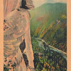 May include: A vintage postcard featuring The Devil's Head at Chimney Rock, Western North Carolina. The image shows a tall, light-colored rock formation overlooking a green valley, a winding river, and U.S. Highway #74, with text at the top and bottom.