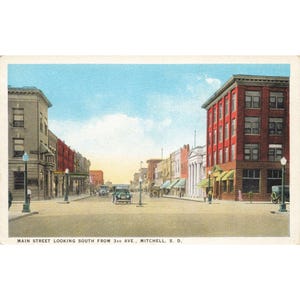 Könnte beinhalten: Vintage Postkarte mit der Main Street in Mitchell, South Dakota, Blickrichtung Süden von der 3rd Avenue. Das Bild zeigt eine Straße mit Gebäuden, Autos und Straßenlaternen unter blauem Himmel mit Wolken. Die Gebäude sind in verschiedenen Farben gehalten, darunter roter Backstein.