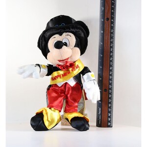 Pode incluir: Um brinquedo de pel&uacute;cia do Mickey Mouse usando uma cartola preta, smoking, cal&ccedil;as vermelhas e faixa amarela com "HAPPY NEW YEAR" impresso. O brinquedo tem aproximadamente 25 cm de altura, com luvas brancas e sapatos amarelos. Uma r&eacute;gua de madeira est&aacute; ao lado do brinquedo.