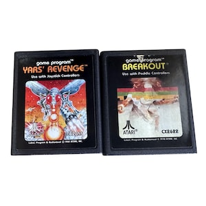 May include: Two Atari game cartridges. The "Yars' Revenge" cartridge features a red and orange graphic of a mechanical insect. The "Breakout" cartridge has a graphic of a person playing a game. Both cartridges are black with white text.