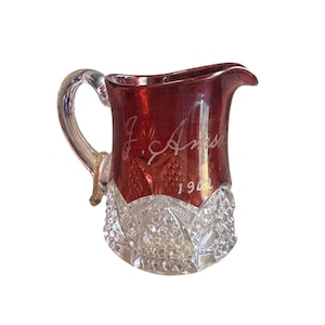 May include: A vintage glass pitcher with a ruby red top and a clear, cut-glass base. The handle is clear glass. Etched on the red portion is the name "J. Amso" and the year "1942".