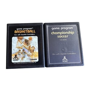May include: Two vintage Atari game cartridges. One is labeled "Basketball" with a basketball player graphic, the other is "Championship Soccer." Both are black with white text and the Atari logo. The Basketball cartridge has the model number CX2624.