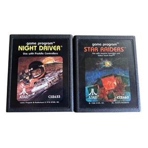 Puede incluir: Dos cartuchos de juego Atari: "Night Driver" y "Star Raiders". "Night Driver" presenta un gráfico de piloto de carreras, mientras que "Star Raiders" muestra una escena de batalla espacial. Ambos cartuchos son negros con el logotipo de Atari y detalles del juego.