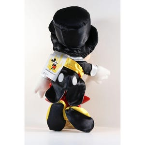Pode incluir: Um brinquedo de pel&uacute;cia do Mickey Mouse usando uma cartola preta, smoking e detalhes amarelos e vermelhos. O brinquedo tem luvas brancas e uma etiqueta com o logotipo da Walt Disney World.