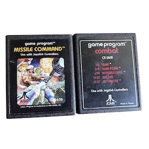 Könnte beinhalten: Zwei Vintage Atari-Spielmodule. Das Modul "Missile Command" zeigt eine Soldatenillustration. Das Modul "Combat" listet Spielmodi auf. Beide Module sind schwarz mit weißem Text und dem Atari-Logo.