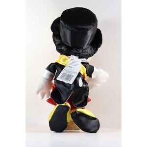 Pode incluir: Um brinquedo de pel&uacute;cia do Mickey Mouse, visto por tr&aacute;s, usando uma cartola preta, jaqueta preta, shorts vermelhos e cachecol amarelo. O brinquedo tem luvas e sapatos brancos. Uma etiqueta diz "NEW YEAR MICKEY BEAN BAG."