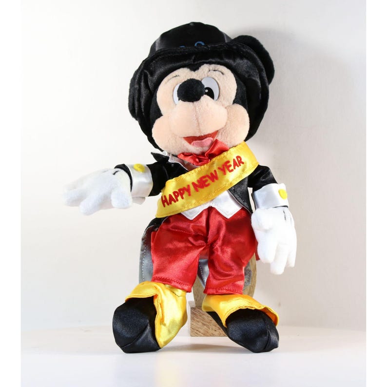 Pode incluir: Uma figura de pel&uacute;cia do Mickey Mouse vestida com um smoking, cal&ccedil;as vermelhas e uma faixa amarela com a inscri&ccedil;&atilde;o "HAPPY NEW YEAR". A figura tem sapatos pretos com detalhes amarelos, luvas brancas e uma cartola preta. A figura est&aacute; sentada.