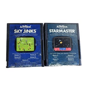 Puede incluir: Dos cartuchos de videojuegos Atari vintage. El cartucho azul de la izquierda es "Sky Jinks" con una pantalla de juego verde y amarilla. El cartucho de la derecha es "StarMaster" con una pantalla de juego con temática espacial. Ambos están etiquetados como "Activision."