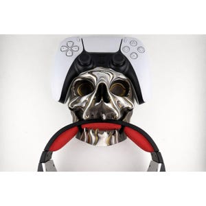 Puede incluir: Una escultura de calavera cromada con un mando de PlayStation 5 blanco y negro encima. Unos auriculares negros y rojos se sujetan en la boca de la calavera. El fondo es blanco liso.
