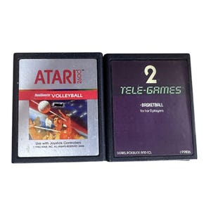 Pode incluir: Dois cartuchos de videogame vintage. Um é rotulado "ATARI 2600 RealSports VOLLEYBALL" com um gráfico de jogador de voleibol. O outro é "TELE-GAMES BASKETBALL for 1 or 2 players". Ambos estão em caixas de plástico preto.