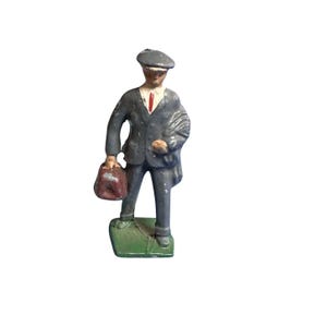May include: A gray lead toy figurine of a man wearing a gray cap, a gray suit, and a red tie. He is holding a brown briefcase in his left hand and is standing on a green base.