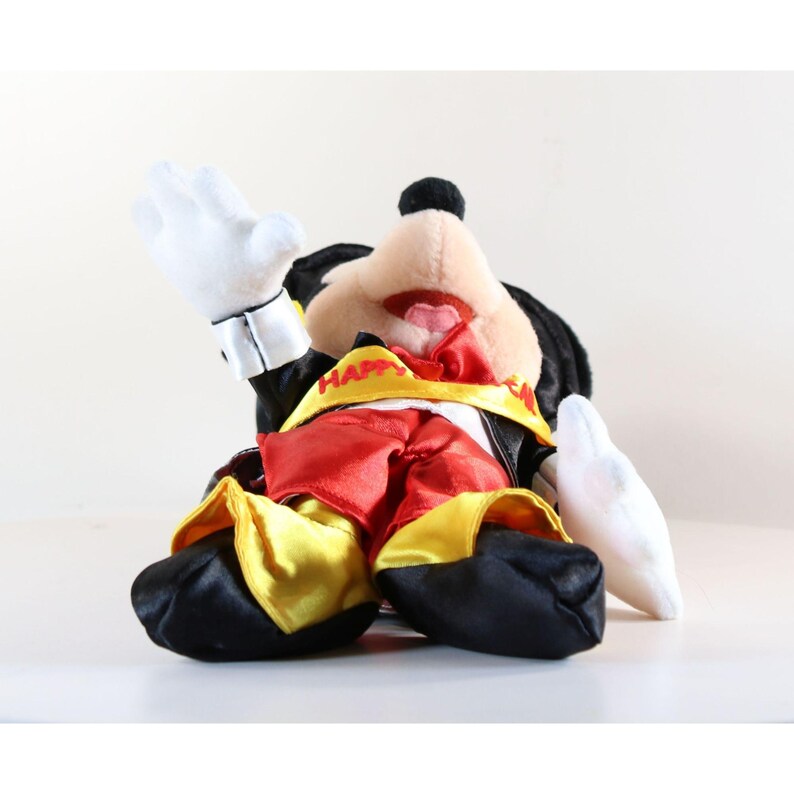 Pode incluir: Boneco de pel&uacute;cia do Mickey Mouse usando uma roupa vermelha e amarela, jaqueta e sapatos pretos. O boneco tem luvas brancas e uma faixa amarela com "HAPPY". O boneco est&aacute; sentado com um bra&ccedil;o levantado.