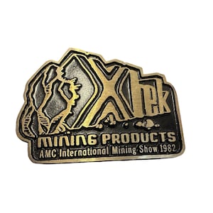 Könnte beinhalten: Eine goldene und schwarze Metallnadel mit dem Text "Xtek Mining Products AMC International Mining Show 1982". Die Nadel zeigt ein stilisiertes Bergpanorama.