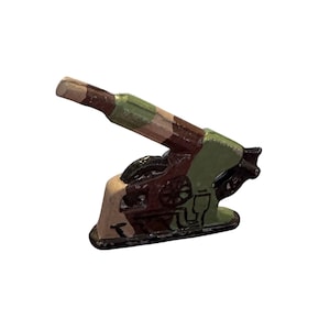 Puede incluir: Un pequeño cañón de juguete pintado en colores de camuflaje verde, marrón y beige. El cañón tiene una rueda y un cañón que apunta hacia arriba.