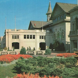 May include: A large stone building with a tall spire and multiple windows stands behind a garden featuring green shrubs, bright red flowers, and a stone fountain. Wooden benches are placed along the stone wall near the building entrance.