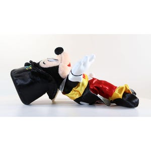 Pode incluir: Um brinquedo de pel&uacute;cia do Mickey Mouse com uma roupa de m&aacute;gico. Mickey est&aacute; deitado de lado, usando uma cartola preta, jaqueta preta, cal&ccedil;as vermelhas e detalhes amarelos e brancos. Suas luvas brancas est&atilde;o levantadas.