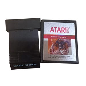 Pode incluir: Dois cartuchos de videogame Atari 2600 pretos. Um é rotulado "SPACE ATTACK" e o outro "RealSports FOOTBALL". O cartucho "FOOTBALL" tem uma ilustração colorida de jogadores de futebol americano.