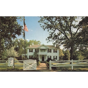 May include: Vintage postcard featuring Murrell Home, a white two-story house with black shutters and a balcony. An American flag waves in the foreground. A white picket fence and gate lead to the house. The sign reads "Murrell Home 1844".