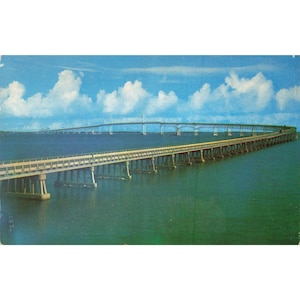 May include: A vintage postcard showing a long bridge stretching across water. The bridge is supported by concrete pillars and has a light-coloured deck. The sky is blue with fluffy white clouds, and the water is a deep teal colour.