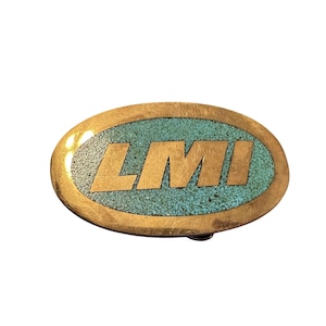 May include: Oval-shaped belt buckle with a gold-colored metal frame and a turquoise inlay. The buckle features the letters "LMI" in gold lettering.