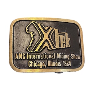 Könnte beinhalten: Rechteckige Metallschnalle mit goldfarbenem Rand und schwarzem Mittelteil. Die Mitte zeigt den Text "Xtek" in Gold sowie den Text "AMC International Mining Show Chicago, Illinois 1984".