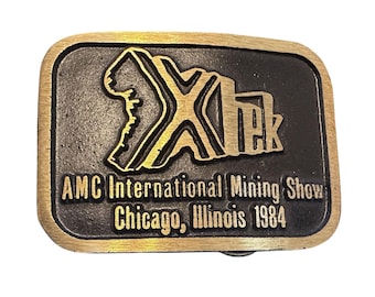 May include: Rectangular metal belt buckle with a gold-colored border and a black center. The center features the text "Xtek" in gold, along with the text "AMC International Mining Show Chicago, Illinois 1984".
