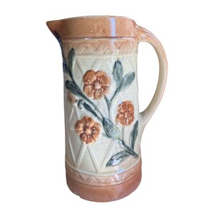 Peut inclure: Pichet en céramique aux tons beige et crème. Il présente un motif floral avec des fleurs orange et des feuilles vertes. La poignée et le bord sont beiges, et le corps a un motif en losange.