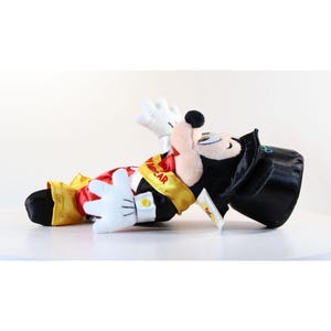 Pode incluir: Um brinquedo de pel&uacute;cia do Mickey Mouse usando uma roupa vermelha e dourada com uma cartola preta. O brinquedo tem luvas brancas e uma faixa amarela que diz "NASCAR". O brinquedo est&aacute; deitado em uma superf&iacute;cie branca.