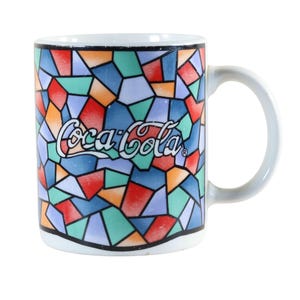 May include: White ceramic Coca-Cola mug with a colorful stained-glass design. The mug features a mosaic pattern of various colors, including red, blue, green, and orange. The Coca-Cola logo is prominently displayed in white.