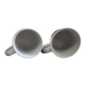 Pu&ograve; includere: Due tazze da caff&egrave; in ceramica bianca con manici curvi sono mostrate su uno sfondo bianco. Le tazze sono vuote e l'interno &egrave; di colore grigio chiaro. I manici sono sul lato destro delle tazze.