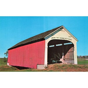 May include: A vintage postcard featuring a red covered bridge under a clear blue sky. The bridge has a white entrance with the text "CROSS THIS BRIDGE AT A WALK". The roof is dark brown, and the bridge is surrounded by green grass.