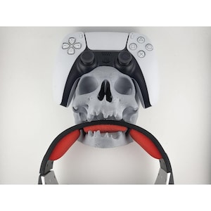Puede incluir: Un soporte gris en forma de calavera para auriculares y mando de juegos. El soporte presenta un mando blanco y negro, una calavera gris y unos auriculares negros con acolchado rojo. Una solución de almacenamiento única para jugadores.