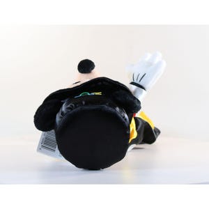 Pode incluir: Um brinquedo de pel&uacute;cia de um personagem de desenho animado, vestindo uma roupa preta com um detalhe amarelo. O brinquedo tem uma cabe&ccedil;a preta, orelhas pretas grandes e uma m&atilde;o enluvada branca levantada em um aceno. O brinquedo est&aacute; deitado de costas.