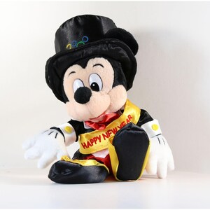 Pode incluir: Um boneco de pel&uacute;cia do Mickey Mouse usando uma cartola preta com an&eacute;is coloridos, uma faixa amarela com o texto "HAPPY NEW YEAR" e uma gravata borboleta vermelha. O boneco tem luvas brancas e sapatos pretos.