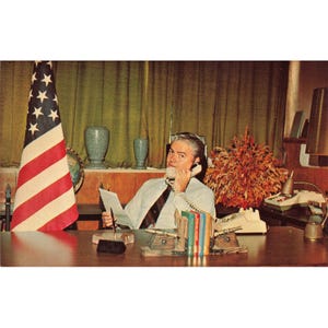 Könnte beinhalten: Vintage-Foto eines Mannes in gestreiftem Hemd und Krawatte, der an einem Schreibtisch sitzt und telefoniert. Links steht eine amerikanische Flagge. Auf dem Schreibtisch befinden sich eine Lampe, Bücher und ein Globus.