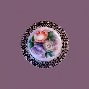 Op de afbeelding: Een ronde broche met een centrale witte porseleinen schijf, handbeschilderd met een boeket van roze, paarse en groene bloemen. De schijf is gevat in een donker, getextureerd metalen frame met een gedraaide koordrand en kleine metalen kraaltjes.
