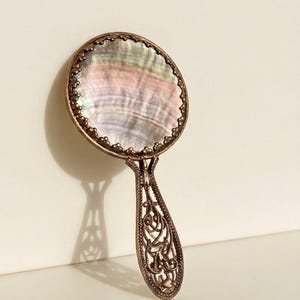 May include: A hand mirror featuring a circular iridescent mother-of-pearl inlay set within a bronze-toned metal frame. The handle displays an intricate openwork filigree design, and the circular border is accented with small, uniform metal beads.