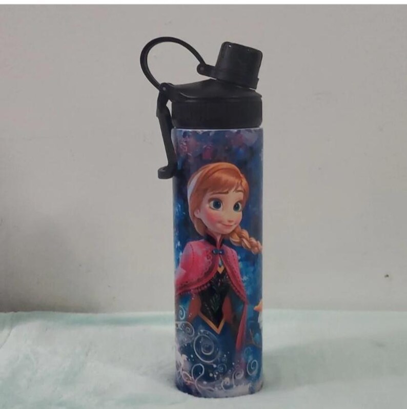 Pu&ograve; includere: Una borraccia cilindrica blu e viola con l'immagine di un cartone animato di una ragazza con i capelli rossi e un mantello rosa. La bottiglia ha un tappo e un manico neri. La bottiglia &egrave; di forma cilindrica.