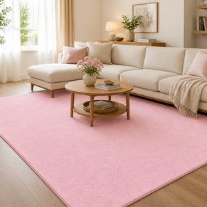 Op de afbeelding: Een groot, rechthoekig, lichtroze tapijt in een woonkamer. Een beige hoekbank en een ronde houten salontafel staan op het tapijt. De kamer heeft houten vloeren en wordt verlicht door natuurlijk licht.