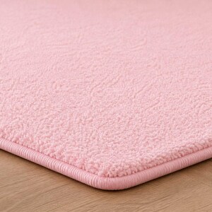 Op de afbeelding: Close-up van een zacht, roze tapijt met een getextureerd oppervlak. Het tapijt heeft een afgebiesde rand in een bijpassende roze kleur. Het tapijt ligt op een lichtbruine houten vloer.