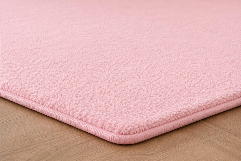 Op de afbeelding: Close-up van een zacht, roze tapijt met een getextureerd oppervlak. Het tapijt heeft een afgebiesde rand in een bijpassende roze kleur. Het tapijt ligt op een lichtbruine houten vloer.