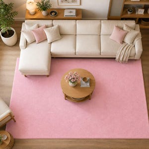 Op de afbeelding: Een bovenaanzicht van een woonkamer met een groot, lichtroze rechthoekig tapijt. Een cr&egrave;mekleurige hoekbank met roze en beige kussens staat op het tapijt. Een ronde houten salontafel en een bijpassende bijzettafel zijn ook aanwezig.