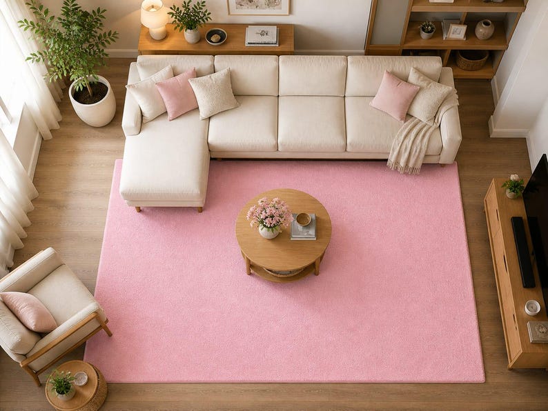 Op de afbeelding: Een bovenaanzicht van een woonkamer met een groot, lichtroze rechthoekig tapijt. Een cr&egrave;mekleurige hoekbank met roze en beige kussens staat op het tapijt. Een ronde houten salontafel en een bijpassende bijzettafel zijn ook aanwezig.