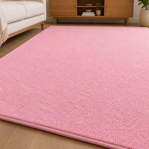 Op de afbeelding: Een groot, rechthoekig roze tapijt met een zacht, getextureerd oppervlak. Het tapijt ligt op een houten vloer en de kleur is effen lichtroze. Het tapijt is een woondecoratie-artikel.