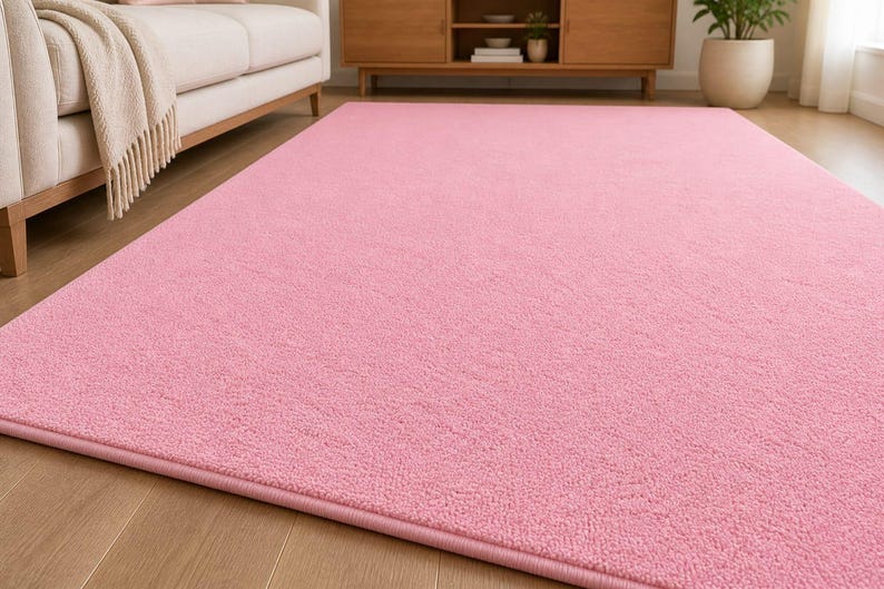 Op de afbeelding: Een groot, rechthoekig roze tapijt met een zacht, getextureerd oppervlak. Het tapijt ligt op een houten vloer en de kleur is effen lichtroze. Het tapijt is een woondecoratie-artikel.